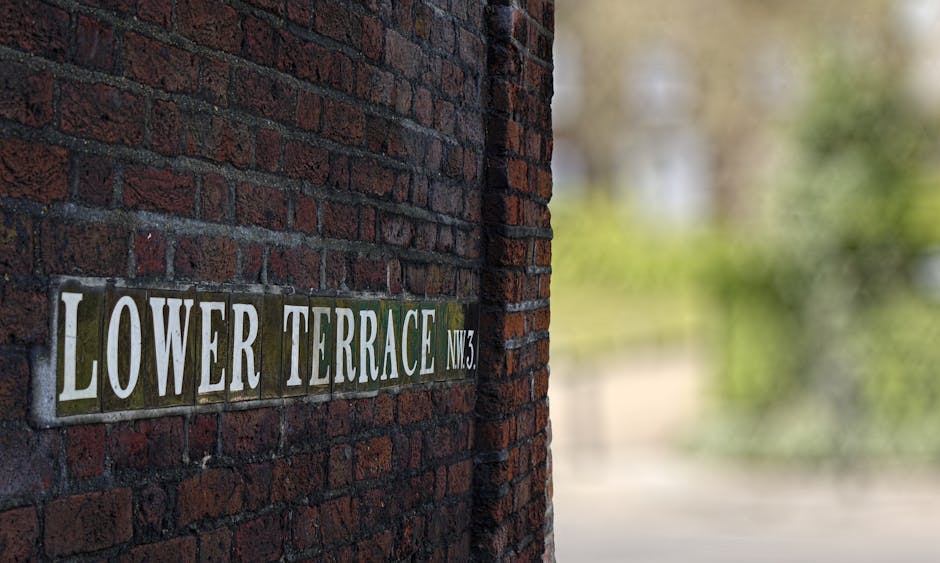 A close-up view of a brick wall with a metal street sign attached, displaying the words 'Lower Terrace NW3' in white lettering on a dark background. The sign is mounted on a red-brown brick surface, which appears clean and well-maintained. In the background, a blurred outdoor scene with greenery and natural light suggests a peaceful residential area in Hampstead, potentially near an exterior wall of a building. The lighting highlights the texture of the bricks and the sign, indicating a bright, sunny day. The overall scene emphasizes the clean and tidy appearance of the outdoor environment, consistent with professional surface cleaning and maintenance practices.