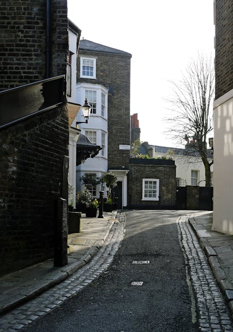 A narrow, paved alleyway in Hampstead lined with traditional brick buildings and residential houses. The pavement features cobblestone edging, and the street is quiet and clean, with a lamppost and potted plants visible near the entrances. The surroundings are bathed in natural daylight, highlighting the well-maintained surfaces and tidy appearance of the area, illustrating a typical scene where professional cleaning services like those from [COMPANY_NAME] could ensure pristine cleanliness and hygiene during end of tenancy cleaning or domestic surface cleaning in the West Hampstead neighborhood.