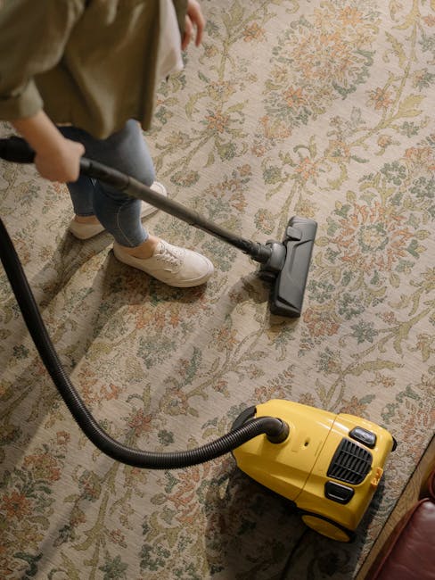 A person using a vacuum cleaner on a patterned carpet in a living room setting. The carpet features floral and geometric motifs in beige, green, and light pink shades. The individual, dressed in casual attire including beige shoes and jeans, is holding the vacuum's handle while the yellow and black vacuum cleaner is positioned on the floor, actively cleaning. The room appears well-lit, with the carpet looking clean and free of visible dust or debris. This scene exemplifies deep cleaning and surface maintenance, typical of residential carpet sanitisation services, with the branding for [COMPANY_NAME] subtly integrated into the context of professional domestic cleaning, as highlighted on the page titled 'Carpet cleaning West Hampstead West End Lane specialists, WEST HAMPSTEAD' at https://carpetcleanerswesthampstead.co.uk.