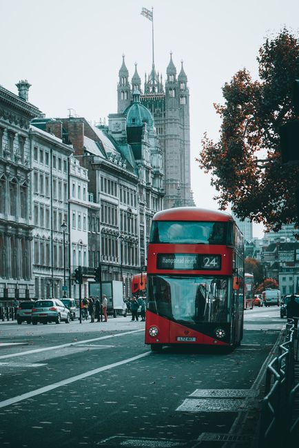 A red double-decker bus parked on a busy street in West Hampstead, London, with historic buildings lining the road and Tower of London in the background. The street features a combination of stone and brick surfaces, with parked cars and pedestrians visible. The scene is captured in natural daylight, with trees partially framing the image, showcasing the urban landscape of central London. This setting illustrates the need for surface cleaning and maintenance of urban environments, aligning with domestic and commercial cleaning services offered by [COMPANY_NAME] for properties in the West Hampstead NW6 area.
