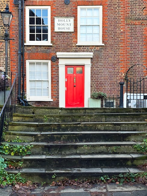 Exterior view of a brick residential building with four white-framed sash windows, one on each floor, and a white sign reading 'Holly Mount House' mounted between the upper windows. The building features a bold red front door with a small window panel and a brass door handle, accessed by a moss-covered stone staircase with five treads. To the right of the door, there is a potted plant and a small green shrub in a planter box, with steps bordered by black metal railings. The scene is lit by natural daylight, showcasing the clean and well-maintained facade, reflecting the professional cleaning and maintenance associated with domestic and surface cleaning services offered by carpetcleanerswesthampstead.co.uk.